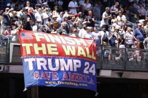 Screaming MAGA fans unfurl a "Finish the Wall" flag at a Trump rally.